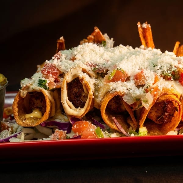 Crispy taquitos topped with cheese, pico de gallo, and a side of guacamole on a red plate.