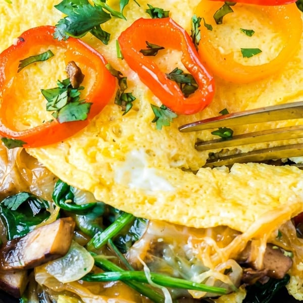 A golden omelet filled with melted cheese, spinach, mushrooms, and bell peppers, with a fork.