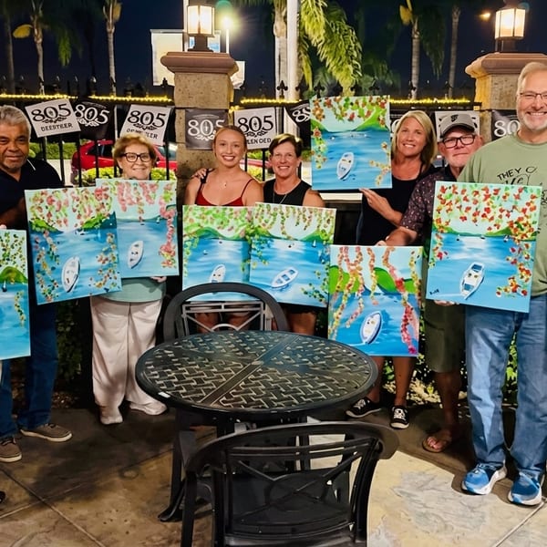 A group of people proudly display their boat and landscape paintings at an outdoor evening event.