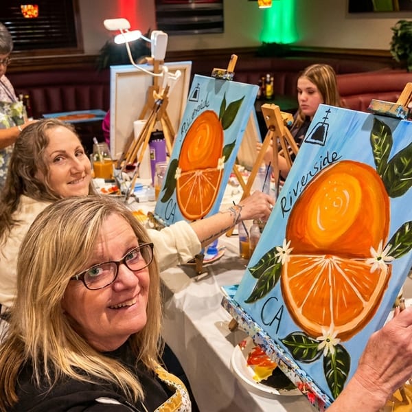 Women painting orange-themed canvases at a restaurant table, smiling at the camera.
