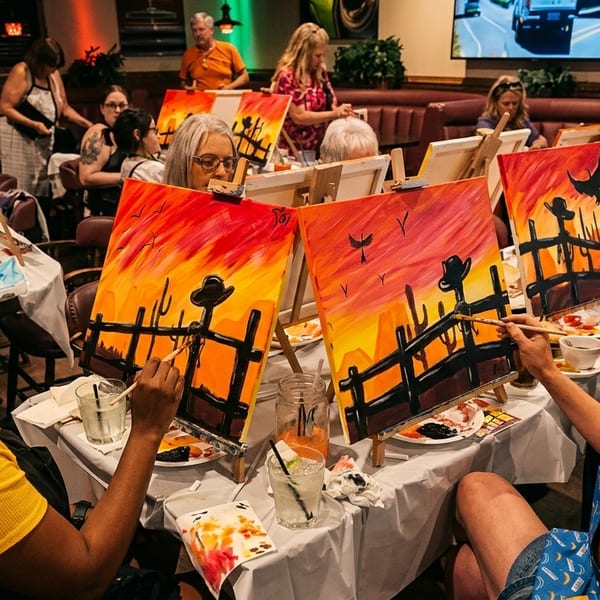People painting desert sunset scenes with cacti and fences at a restaurant event.