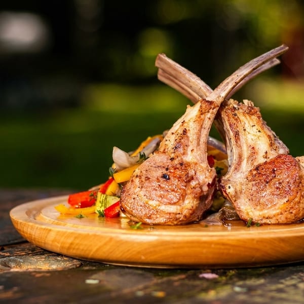 Two grilled lamb chops with colorful vegetables on a wooden plate, set on a rustic outdoor table.